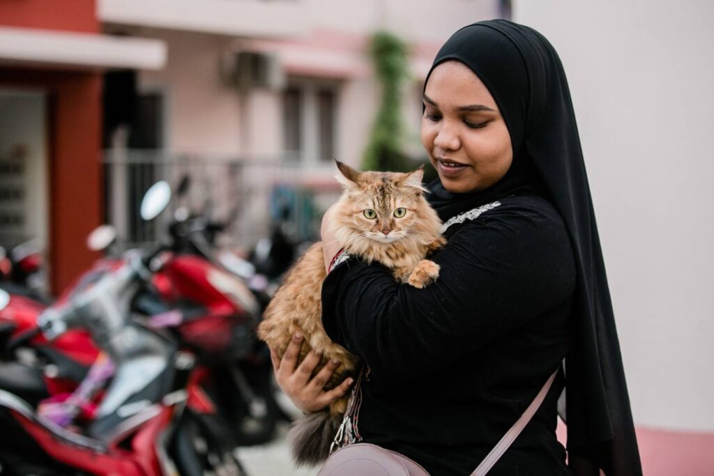A smiling woman in a hijab holds a tabby cat outdoors